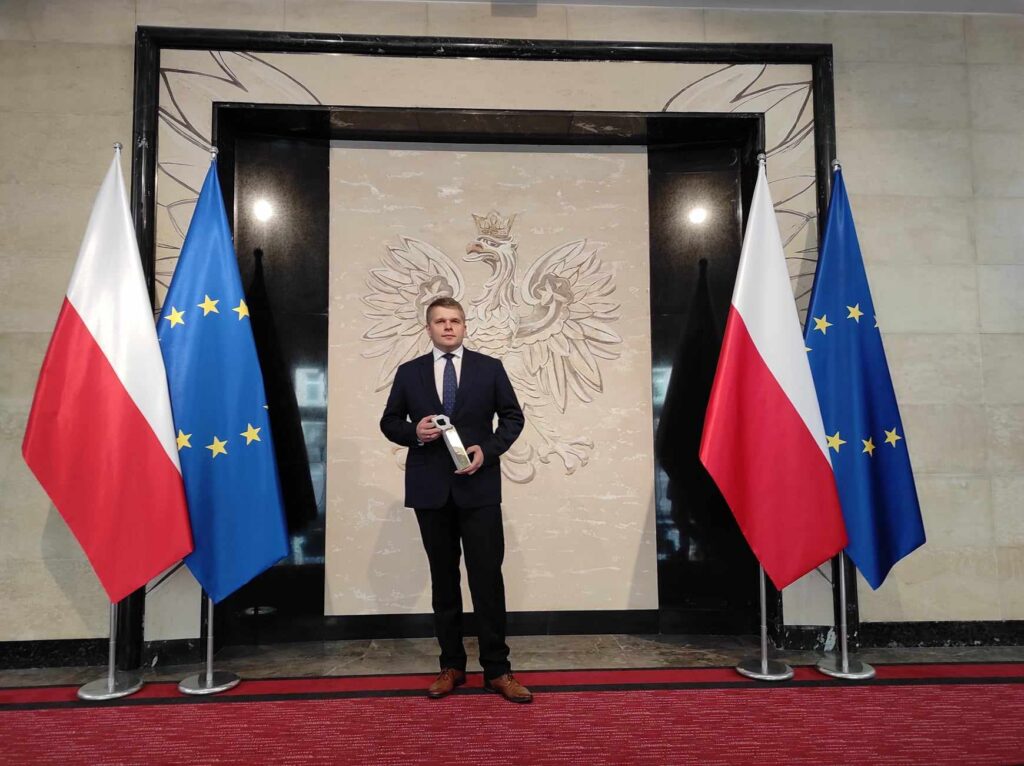 Zdjęcie mgr inż. Łukasza Ślaga na tle ściany z godłem Polski i flagami Polski i Unii Europejskiej